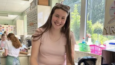 A mid shot of Vene. She has long brown hair, is smiling and wearing a light pink top. She is standing over the kitchen worktop.