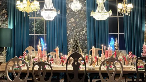 IWC Media Elegant dining room with a long table set for Christmas, featuring candles, miniature trees, and festive decorations under chandeliers.