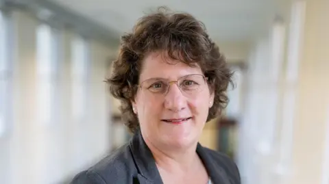 Queen Elizabeth Hospital Chief executive Alice Webster wears a dark grey suit and a white patterned top. She is standing in a hospital corridor. 