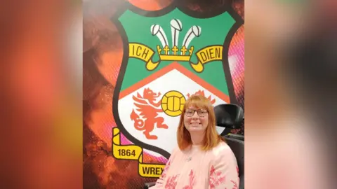 Kerry Evans Kerry Evans, pictured in her wheelchair in front of Wrexham FC's football badge. She wears a pink floral patterned blouse and smiles with her teeth at the camera. She has ginger bobbed hair and wears tortoiseshell square glasses. 