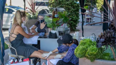 Los Angeles Times via Getty Images Beauty technicians wear face masks as they give a client wearing a face mask a manicure and pedicure outdoors