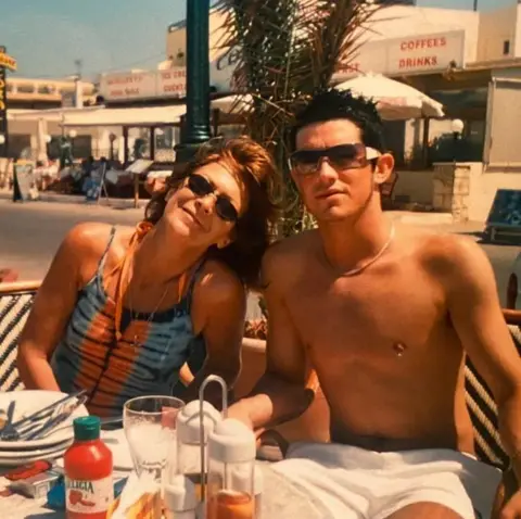 A woman in sunglasses sitting with a younger man in white shorts in a sunny overseas looking place