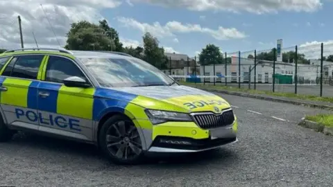 A police car is seen beside a school that has a cordon. The school fence is seen outside a large white building.