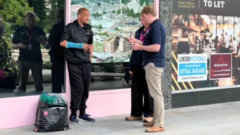A man, who is bald and wearing a blue arm cast and black clothing, stands outside a shop while two council officers speak to him. There are a couple of shopping bags containing sleeping bags beside him.