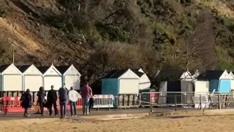 Six beachgoers watch as part of a cliff collapses onto beach huts