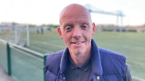 Andy Campbell smiling at the camera. He is wearing a grey long-sleeved shirt and a blue body warmer. He is bald. The football pitch is behind him with Middlesbrough's Transporter Bridge in the distance