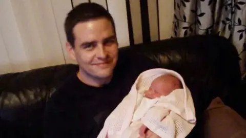 Christian Bagley is sat on a black sofa smiling while holding a baby wrapped in white blankets. He has dark spikey hair and is wearing a black jumper.