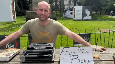 Patrick, a man with a shaved head and stubbled facial hair, smiles at the camera. He is sat on a wooden bench with a typewriter and a sign reading 'free typewriter poems' and is sitting in front of a patch of green.