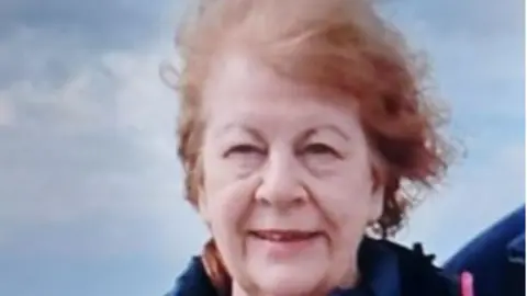 Norfolk Police Close-up of Mary White smiling at the camera. She has red hair, which is now grey, and a blue jacket. The sky fills the background.