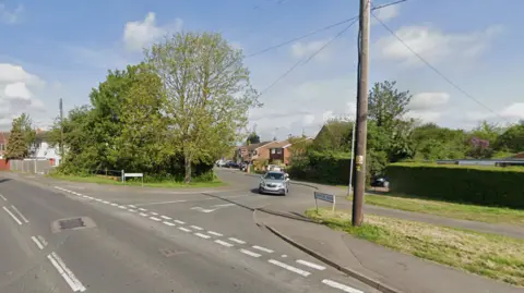 A grey car driving towards us from a junction onto a main road. Beside the junction is a footpath, a patch of grass and a brown wooden phone pole. The sky is blue and cloudy