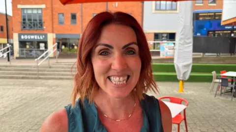 Ruth Davies smiles for the camera. She is around 40-50 years old with neck-length dyed red hair. She's wearing a small necklace and a turquoise top. Cafe tables and chairs can be seen in the background