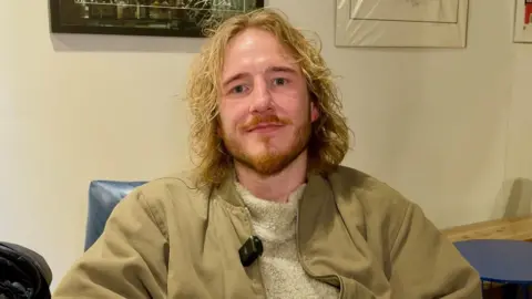 Alex has shoulder-length blonde hair and a ginger beard. He is wearing a khaki coloured bomber jacket and fluffy cream jumper. He is sitting in an armchair with artwork behind him on the walls.