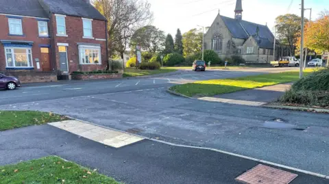 The junction at the A690 and St John's Road at Meadowfield, as pictured from the entrance road to council offices opposite. The main A690 is a narrow two-lane road lined with terraced houses on one side of the St John's Road junction and a church in mature grounds on the other. A car is turning into St John's Road while a flat-bed lorry appears to have just turned out of it. 