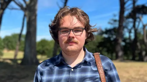 Charlie Roberts is a young man with brown hair, wearing glasses, a blue checked shirt and brown bag strap. He is looking at the camera.
