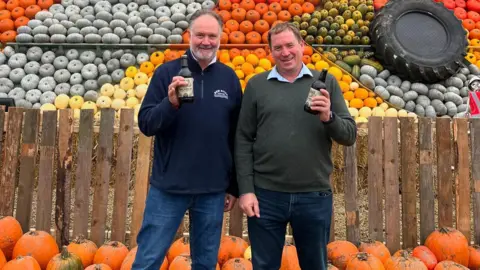 Cammas Hall Farm Jonathan Lukies stands next to another man, they are both looking directly at the camera and holding up beers in front of the mosaic. They are both wearing jeans and dark-coloured jumpers. Rows of pumpkins can be seen behind them at their feet. 