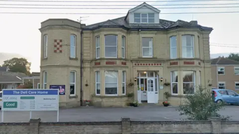 An Edwardian-looking yellow-brick large detached building. It has bow windows and a mansard window in the roof. In front of it is a car park and a low wall and a sign on the left saying The Dell Care Home. 