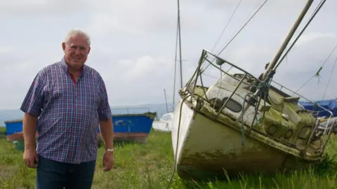 Councillor Andrew Hodson has white cropped hair and is wearing a red, white and blue checked short-sleeved shirt and blue trousers. He is standing next to a rusty boat and in front of a rotten blue one.
