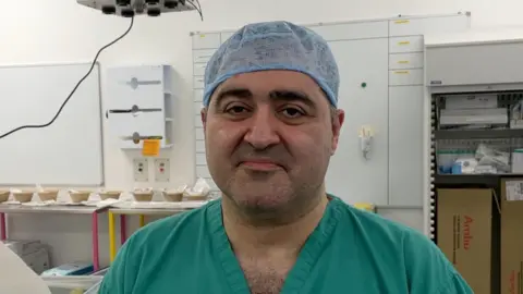 Prof Reza Nouraei, is smiling at the camera. He has dark brown eyes and eye brows. He is wearing a green hospital scrub with v neck. He has a blue hair net on. He is in a hospital setting, behind him are hospital white boards.