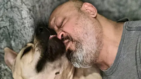 A bald man in a grey t-shirt with a grey beard kisses his dog while lying down together. The dog has a black nose and cream coloured fur.