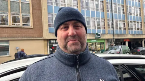 Mr Saker, pictured on a busy street standing next to a parked car. He is wearing a dark blue/grey fleece, zipped up to the top, and a dark blue beanie. 