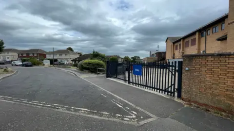 Laura Foster/BBC An area in Walton-on-the-Naze in Essex where an attack on an 82-year-old man took place. The area has a road and brown bricked buildings. There is also a black fence with a blue rectangular sign on it which says Entry. 