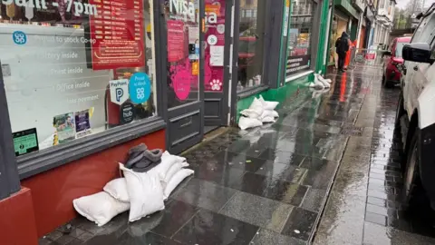 Stacks of white sandbags are placed on the street which is wet from rain. The shop in the forefront is a Nisa convenience store. 