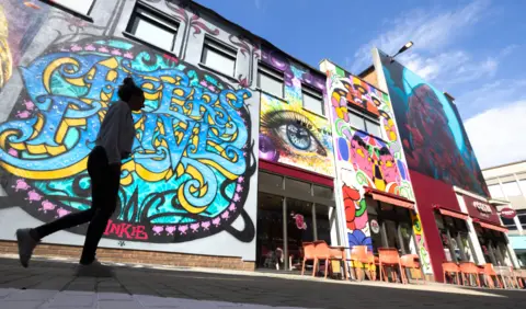 Paul Box Various art work by street artists on a Bristol street, including a 'cheers drive' slogan in blue and yellow, a beautiful eye and a love saves the day wall in rainbow colours. There is a Costa Coffee shop to the right of the picture with a picture of ALfred the gorilla above it.