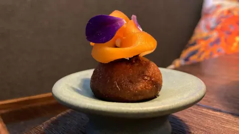 An image of a fried carrot donut with shaved carrot on top, and a decorative flower.