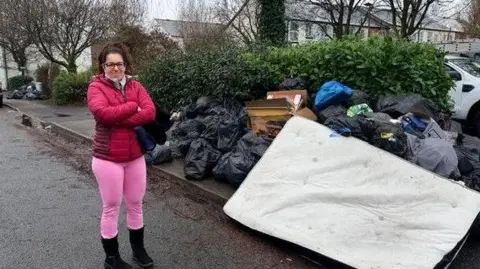 Gabriel Bononi A woman in a dark pink jacket, bright pink leggings and black boots stands in the road next to a pile of a black bin bags and boxes. There is a mattress added to it too