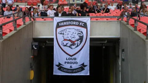 Getty Images A white flag with a bull illustrated in black printed on it. The words "HEREFORD FC, loud proud and forever united" are printed on the flag. There are people sat above the flag in the football stands that have red plastic chairs