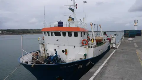 The Gry Maritha docked. It is a small white boat with room for cargo to be stored on. It has an orange strip on it. In the background there is some land visible and a large body of water.