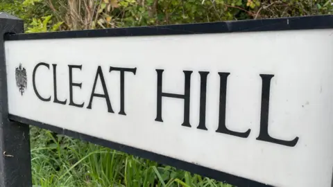 Helen Mulroy/BBC A close-up of a white road sign saying Cleat Hill in black block capitals. Behind it is grass and foliage.
