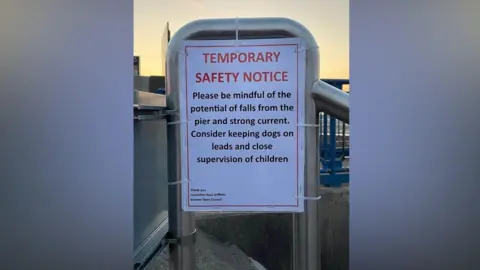 A laminated paper signage attached to a ramp reads TEMPORARY SAFETY NOTICE and advises people to be mindful of potential falls and keep dogs on leads. 