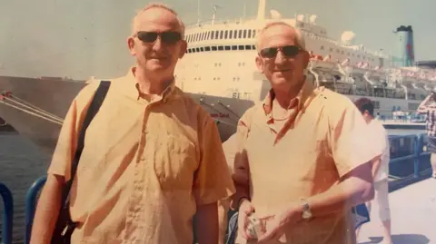 The twins are posing infront of a cruise ship on the water. They both are wearing sunglasses and have yellow button up shirts. They are smiling slightly at the camera. 