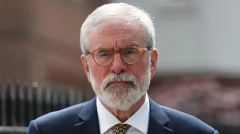 Gerry Adams is on a street. He has white hair and a beard. He's wearing orange glasses and a navy suit with a white shirt and a tie.