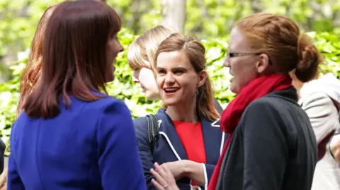 PA Media Image shows a woman in a red top and blue jacket faced towards the camera. 
She is speaking to two women, one in a bright blue jacket with her back to the camera, the other in a red scarf with a navy coat also turned away from the camera. 