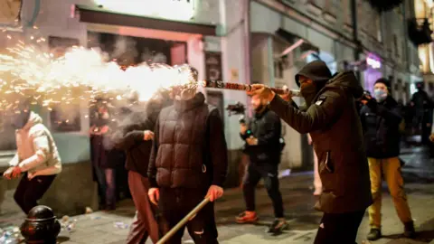 EPA A Georgian opposition supporter in a jacket launches a firework towards police. He is wearing a hood and a mask and is surrounded by other protesters