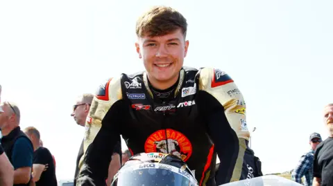 Storm Stacey sitting on his motorbike at a race weekend. He is smiling and wearing a black motorbike suit. 