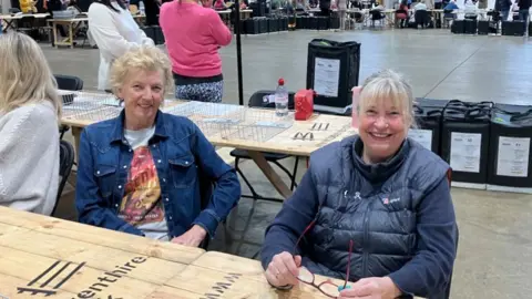 Alex Macdonald and Christine Wilson, who helped to count votes, sit at the end of a long table in a large hall. they are both wearing blue jackets.