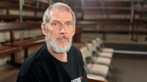 Keith Irons is a man with grey hair and a grey beard. He is wearing a dark blue or black top and behind him there are rows of chairs, out of focus.