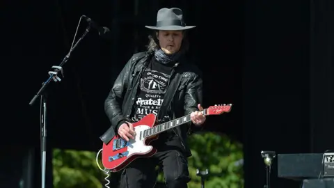 Jon Fratelli onstage - he is playing guitar, wearing a black hat, black T-shirt and leather jacket