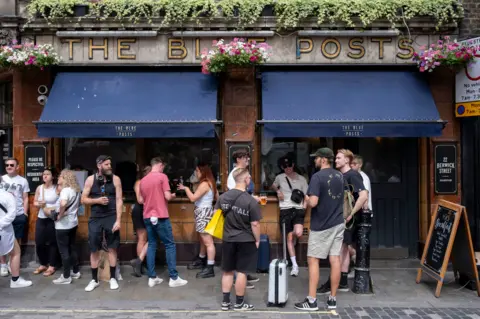 AFP via Getty Images A group of people stand on the pavement drinking outside The Blue Posts pub in London. The pub has blue awnings over the windows with The Blue Posts written in gold above them