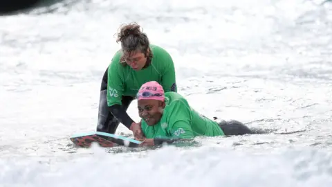 The Wave A woman lying on a bodyboard at The Wave in Bristol. She is wearing a wetsuit, green rash vest and pink swimming cap. A female volunteer is standing beside her and holding her shoulder, ready to guide her into catching a wave.