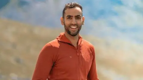 PA Media Sameed Asghar is standing in front of a hillside which is blurred in the background. He has short dark hair and dark facial hair. He is wearing a orange top