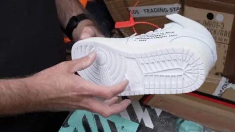 Dan is holding a white trainer which has black text and an orange tag on it.