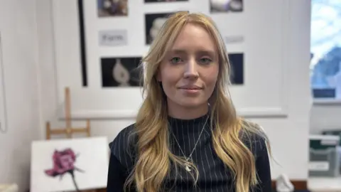 A female art teacher with long blonde hair, wearing black, in a classroom 