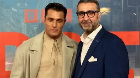 A man in a light-coloured coat and shirt standing next to another man in glasses and a blue suit jacket