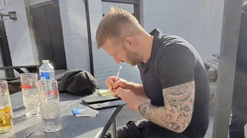 Counter Terrorism Policing A man with blond hair and beard and a tattooed arm, dressed in a black polo shirt and black cargo pants, sits at a table writing notes. He is looking at a phone. 