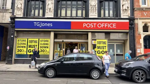 The front of a Post Office and TG Jones shop on a High Street. Cars are parked outside on the road and people are walking past as well as in and out of the shop.