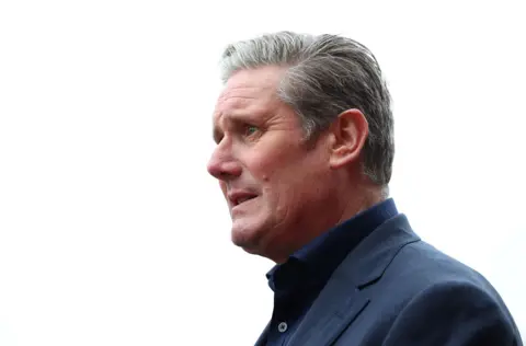 Prime Minister Sir Kier Starmer is seen in a side profile shot against a white background. He is wearing a dark blue jacket and dark blue shirt with no tie